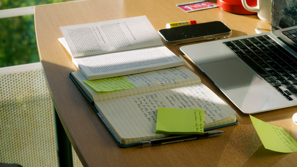 Libro, cuaderno con apuntes y ordenador sobre una mesa junto a una ventana.